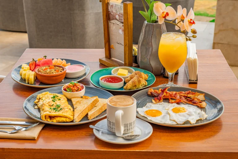 Desayuno completo con huevos, tocino, fruta, pan y café; jugo natural en la mesa